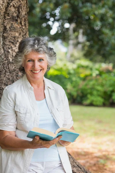 Lächelnde reife Frau liest Buch angelehnt an Baumstamm — Stockfoto