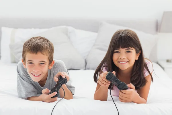 Irmãos sorridentes deitados na cama jogando videogames juntos — Fotografia de Stock