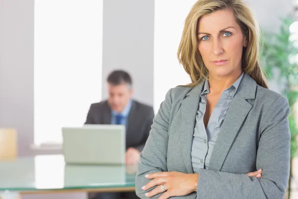 Lächelnde Geschäftsfrau, die in die Kamera blickt, verschränkte Arme mit einem Geschäftsmann, der im Hintergrund arbeitet — Stockfoto