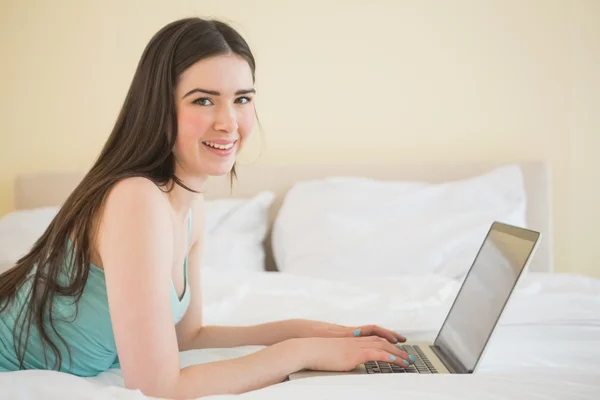 Chica contenta mirando a la cámara usando un portátil acostado en una cama —  Fotos de Stock