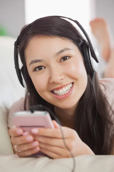 Весела азіатська дівчина лежить на дивані і слухає музику зі смартфоном, дивлячись на камеру — стокове фото