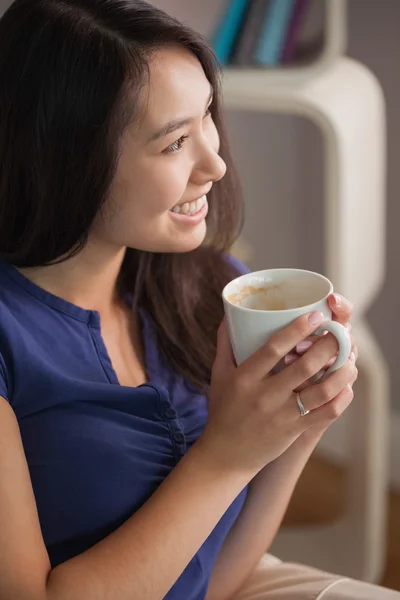 커피는 멀리 보는 낯 짝을 들고 소파에 앉아 행복 한 아시아 여자 — 스톡 사진