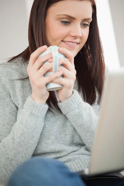 Smiling girl sitting on a sofa holding a cup of coffee and looking at laptop — Stock Photo, Image