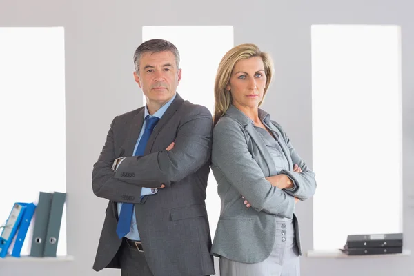 Two unsmiling businesspeople looking at camera standing back to back with crossed arms — Stock Photo, Image