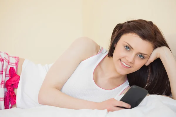 Smiling girl looking at camera using her phone lying on a bed — Stock Photo, Image