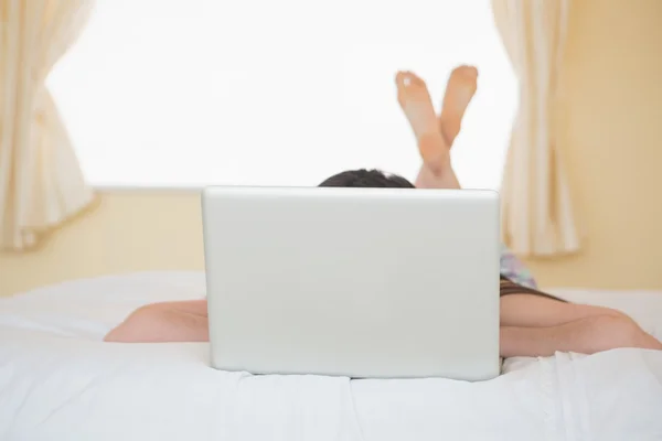 Chica acostada en una cama escondida detrás de un ordenador portátil —  Fotos de Stock