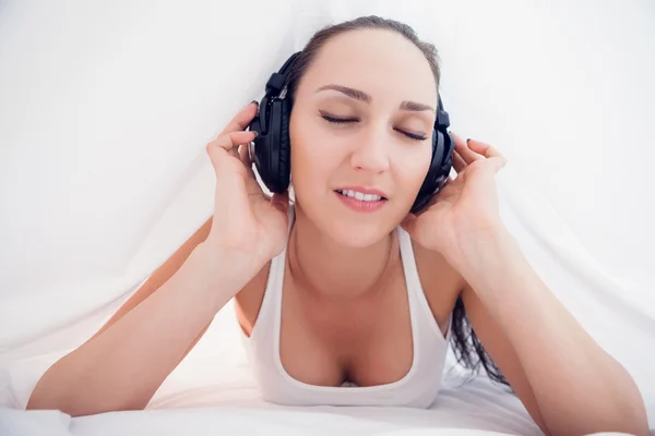 Happy brunette lying under the sheets listening to music — Stock Photo, Image