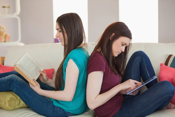 Två glada vänner sitter rygg mot rygg på soffa läsa boken och använder tablet — Stockfoto