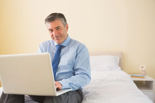 Uomo sorridente che utilizza un computer portatile seduto su un letto — Foto Stock