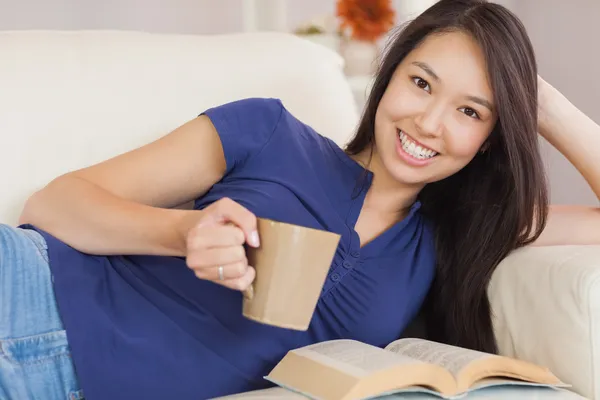 Привлекательная молодая азиатка лежит на диване, читает роман и пьет горячий напиток. — стоковое фото