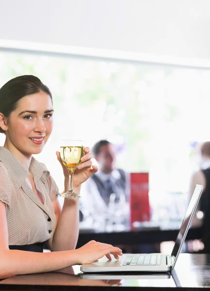 Mujer de negocios sonriente usando el ordenador portátil mientras sostiene la copa de vino —  Fotos de Stock