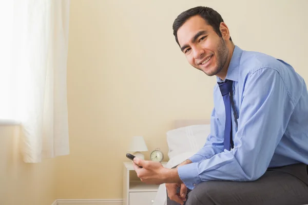 Hombre agradable mirando la cámara sentada en una cama usando un teléfono móvil — Foto de Stock