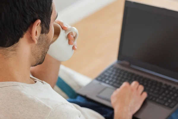 Bonito homem de cabelos castanhos bebendo café enquanto usa seu laptop — Fotografia de Stock