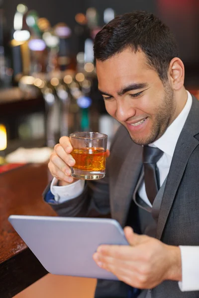 Улыбающийся бизнесмен работает над своим планшетом, выпивая виски — стоковое фото