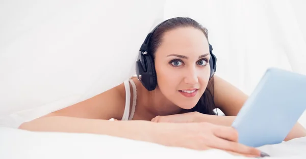 Szczęśliwy brunette leżącego w arkuszach, słuchanie muzyki za pomocą jej komputera typu tablet — Zdjęcie stockowe