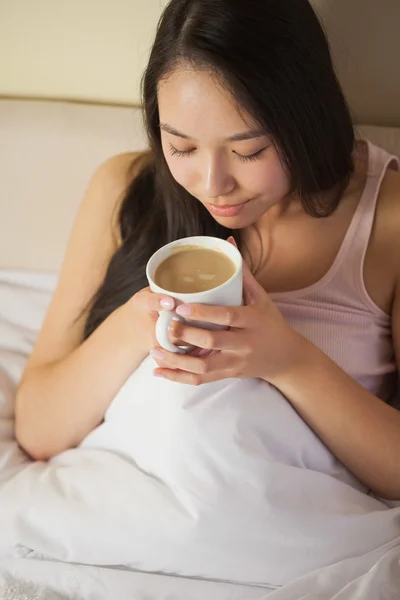 Cheerful young asian woman sitting in bed smelling her morning coffee — Stock Photo, Image