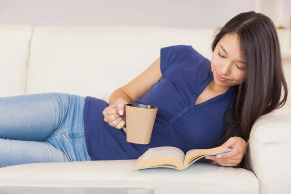 Hermosa chica asiática tumbada en el sofá leyendo una novela y bebiendo bebida caliente — Foto de Stock