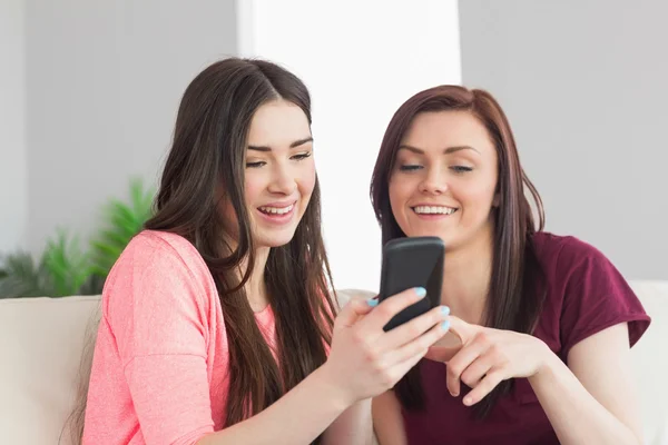 Две улыбающиеся девушки сидят на диване и печатают на мобильном телефоне — стоковое фото