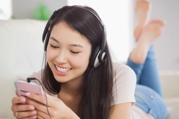 Menina asiática feliz deitado no sofá e ouvir música com smartphone — Fotografia de Stock