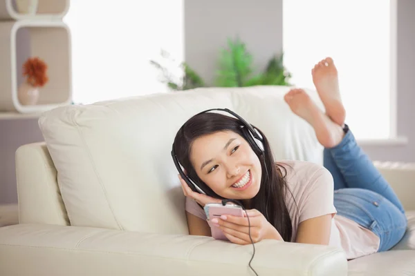 Usmívající se Asijská dívka leží na pohovce a poslouchá hudbu s smartphone — Stock fotografie