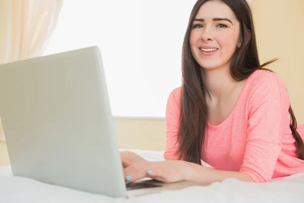 Pretty young girl looking at camera using a laptop lying on a bed — Stock Photo, Image