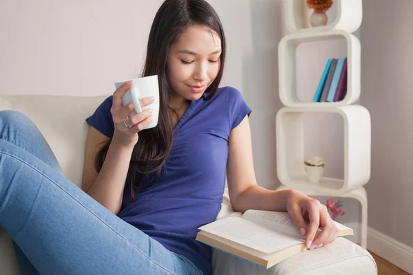 Lectura joven asiático mujer sentado en el sofá celebración taza — Foto de Stock