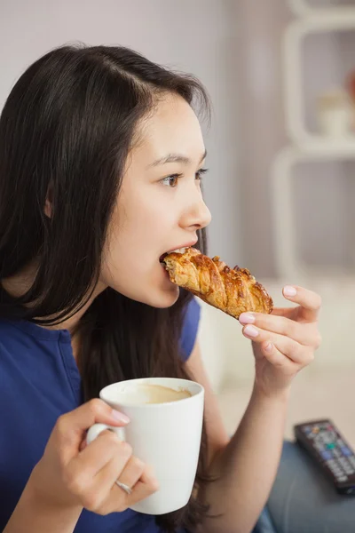 Молодая азиатка, сидящая на диване, пьет кофе и ест выпечку. — стоковое фото