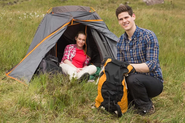 Улыбающийся мужчина собирает рюкзак, пока девушка сидит в палатке — стоковое фото
