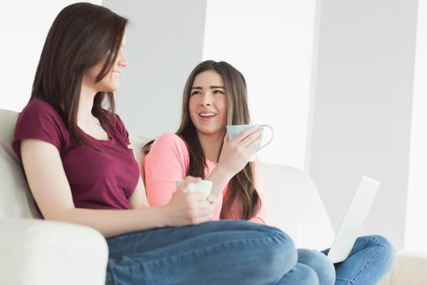 Twee lachende meisjes zittend op een bank u geniet van een drankje en houden van een laptop — Stockfoto