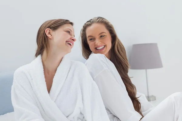 Happy friends wearing bathrobe sitting on bed — Stock Photo, Image