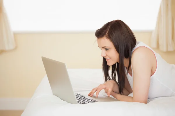 Chica alegre usando un ordenador portátil acostado en su cama —  Fotos de Stock