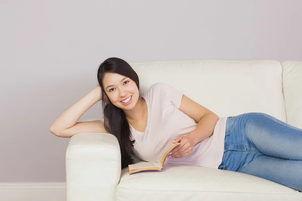 Feliz chica asiática acostada en el sofá leyendo libro sonriendo a la cámara — Foto de Stock