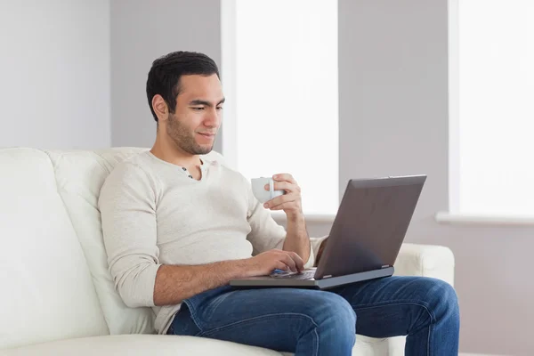 Entspannter attraktiver Mann hält Kaffee, während er an seinem Laptop arbeitet — Stockfoto