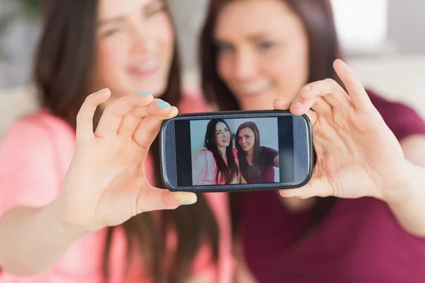 Two happy girls sitting on a sofa taking a photo of themselves with a mobile phone — Stock Photo, Image