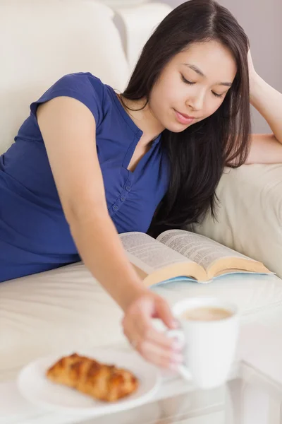 Hezká mladá asijská žena ležící na pohovce, čtení knihy sáhl pro kávu — Stock fotografie