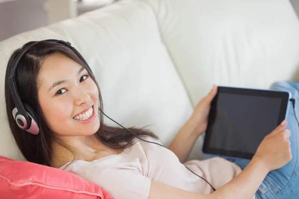 Chica usando su tableta PC en el sofá y escuchando música sonriendo a la cámara —  Fotos de Stock