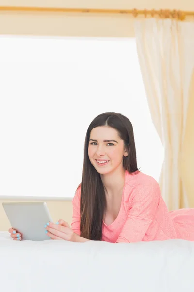 Pretty smiling girl looking at camera and using a tablet pc laying on a bed — Stock Photo, Image