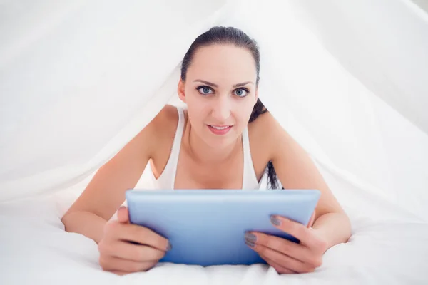 Szczęśliwy brunette leżącego w arkuszach, trzymając jej komputera typu tablet — Zdjęcie stockowe