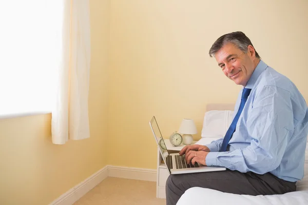 Hombre agradable usando un ordenador portátil sentado en una cama — Foto de Stock