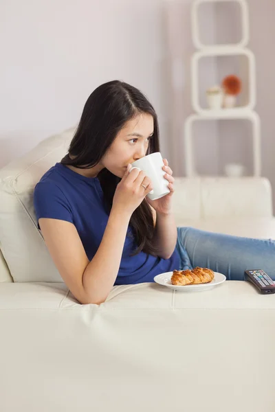 Woman sitting on the sofa drinking coffee with a pastry — Stock Photo, Image