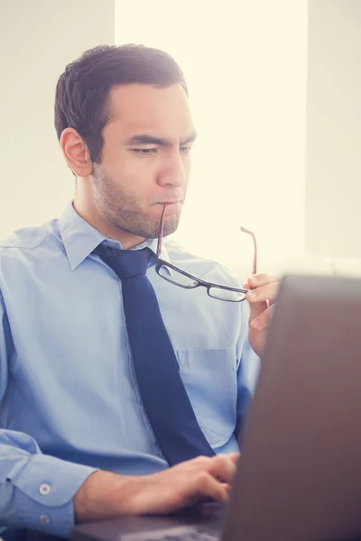 Focused man biting his eyeglasses and using a laptop — Stock Photo, Image