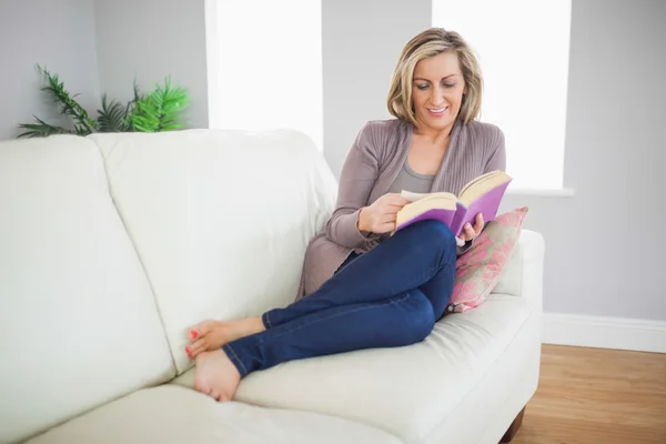 Lächelnde Frau, die auf einem Sofa liegt und ein Buch liest — Stockfoto