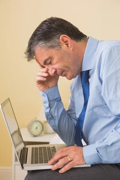 Tired man using a laptop sitting on a bed — Stock Photo, Image