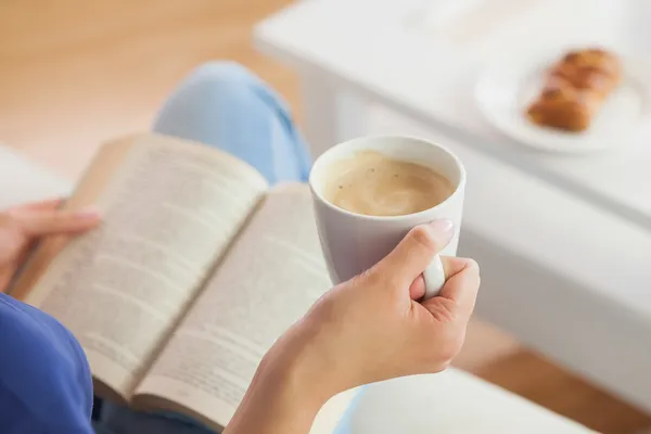 Wanita duduk di sofa membaca buku memegang cangkir kopinya Stok Foto
