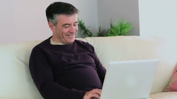 Улыбающийся мужчина использует ноутбук на диване, а жена садится рядом с ним. — стоковое видео