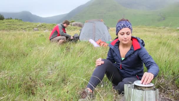 Женщина готовит на открытом воздухе, пока бойфренд ставит палатку — стоковое видео