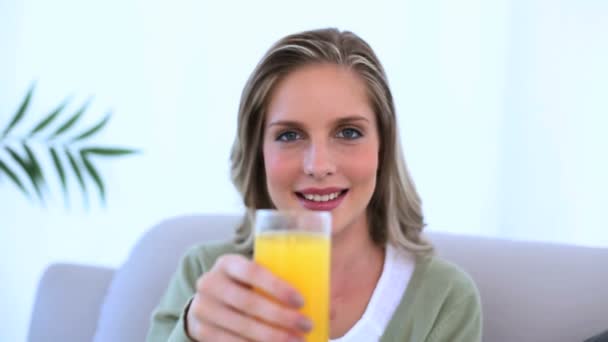 Mujer sentada en el sofá mostrando su vaso de jugo de naranja — Vídeos de Stock