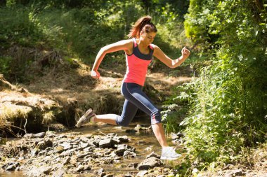 Sporty young woman leaping over a stream clipart