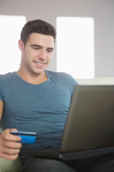 Χαμογελώντας όμορφος άντρας χαλαρωτικό στον καναπέ, χρησιμοποιώντας φορητό υπολογιστή ψώνια σε απευθείας σύνδεση — Φωτογραφία Αρχείου