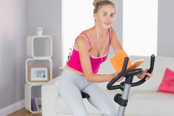 Sporty smiling blonde training on exercise bike reading a book — Stock Photo, Image
